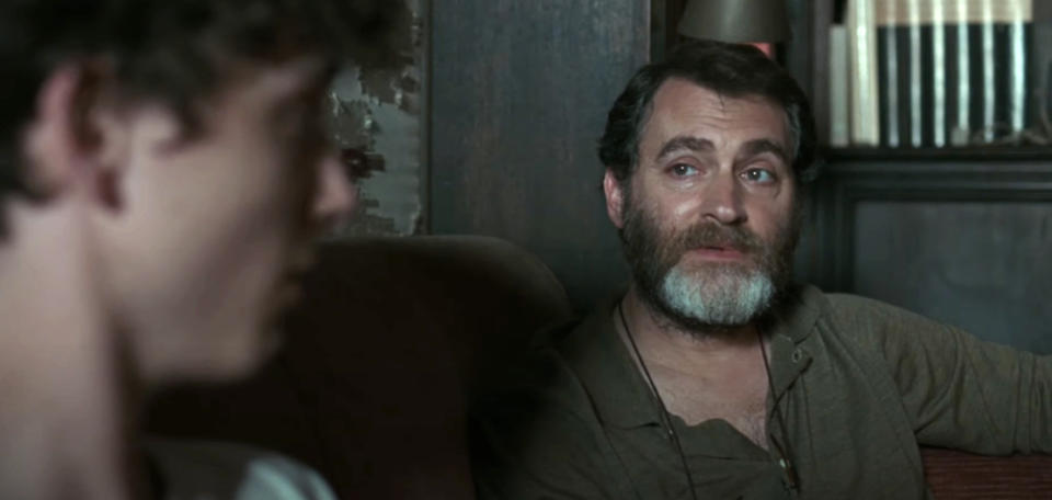 Two characters, one younger and one older with a beard, having a serious conversation in a cozy, dimly-lit room filled with bookshelves