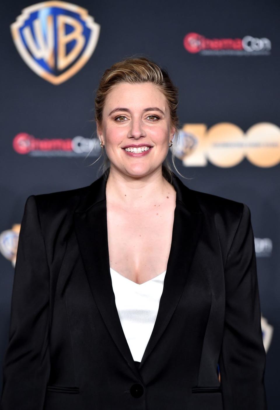 greta gerwig smiles at the camera, she wears a black suit jacket and white top and stands in front of a blurry black background with logos