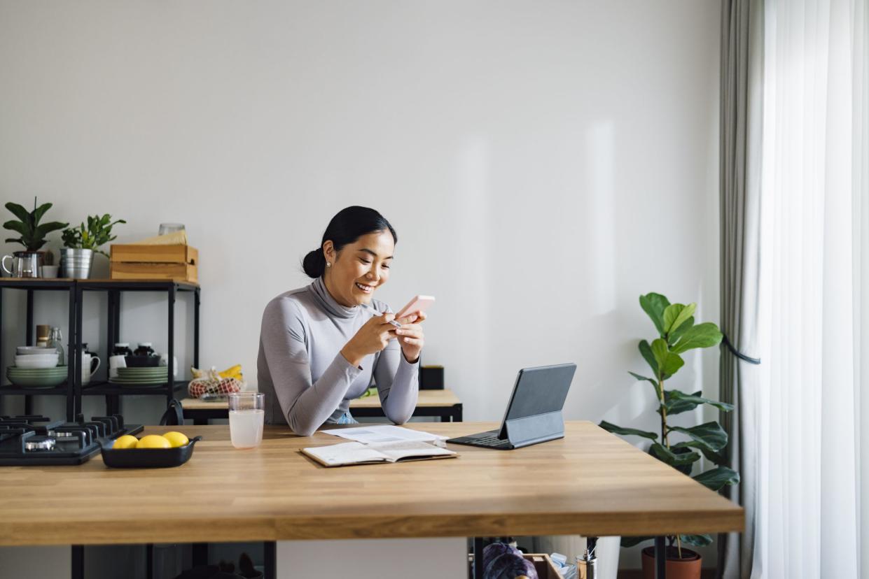 Beautiful Asian businesswoman using digital tablet and notebook while typing text message on her mobile phone at kitchen desk.