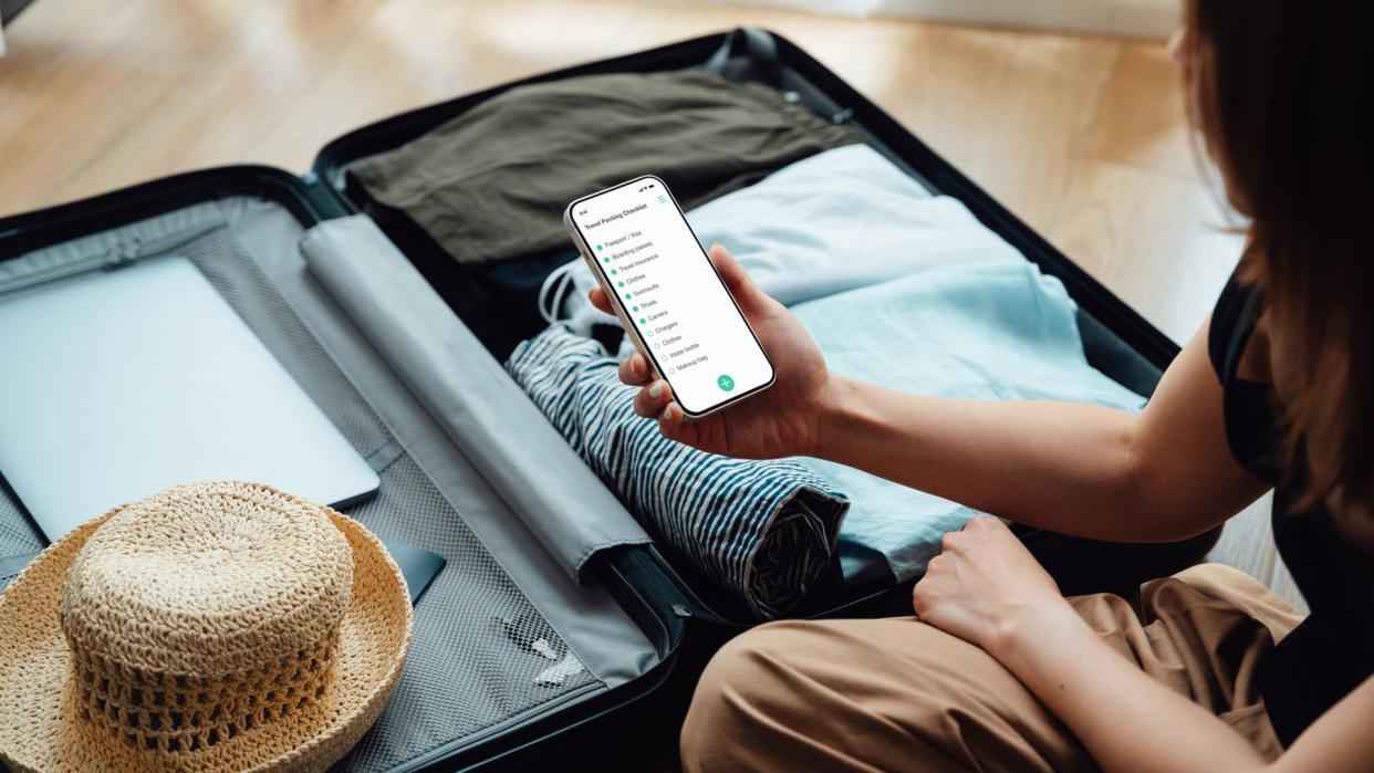 Business woman using smartphone while packing suitcase, getting ready for a work trip. Business and leisure travel.