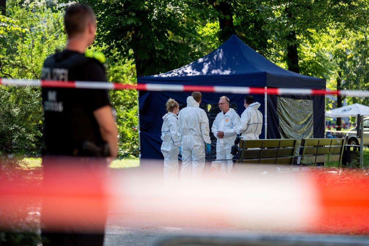 Several officials in white hazmat jumpsuits confer by a temporary canopy by some park benches fenced off by orange and white crime tape, as a security guard looks on.