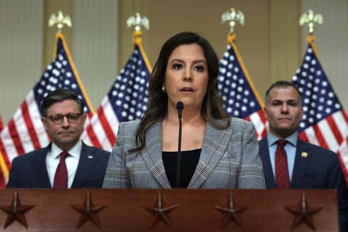 Woman at podium with microphone, flanked by two men, in front of US flags