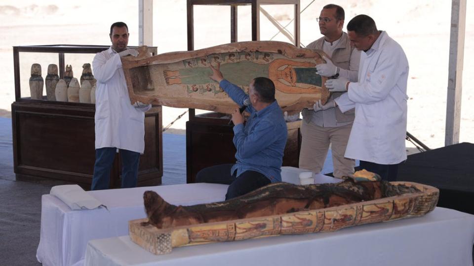 A speaker points to decorations inside the lid of a sarcophagus, which is being held up by three people.