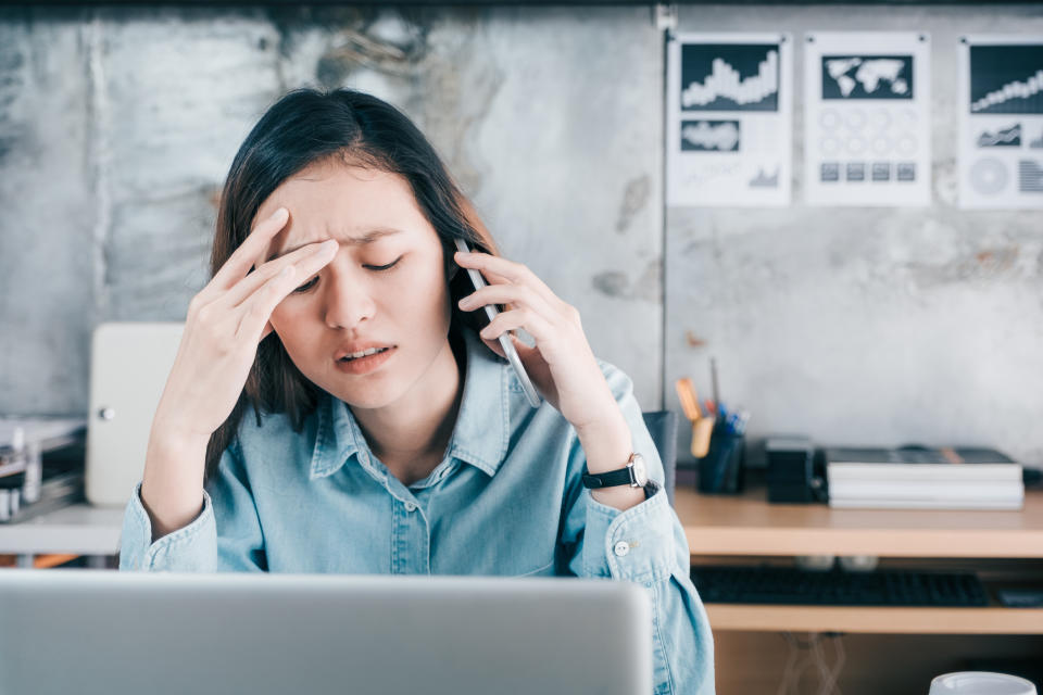 Stressed woman covers her face with hand and feels upset while talking on mobile phone in front of laptop computer at office.