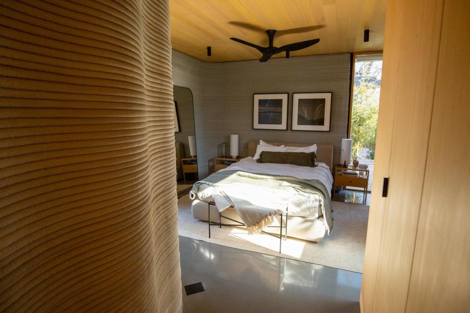 Inside Icon's over 2,000-square-foot House Zero in Austin. There's a bedroom with a bed, mirror and ceiling fan in front of a layered printed concrete wall.