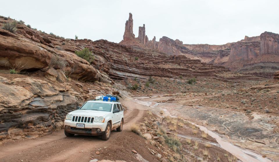 An SUV drives past Monster Tower and Washer Woman Arch along White Rim Road in Canyonlands National Park.