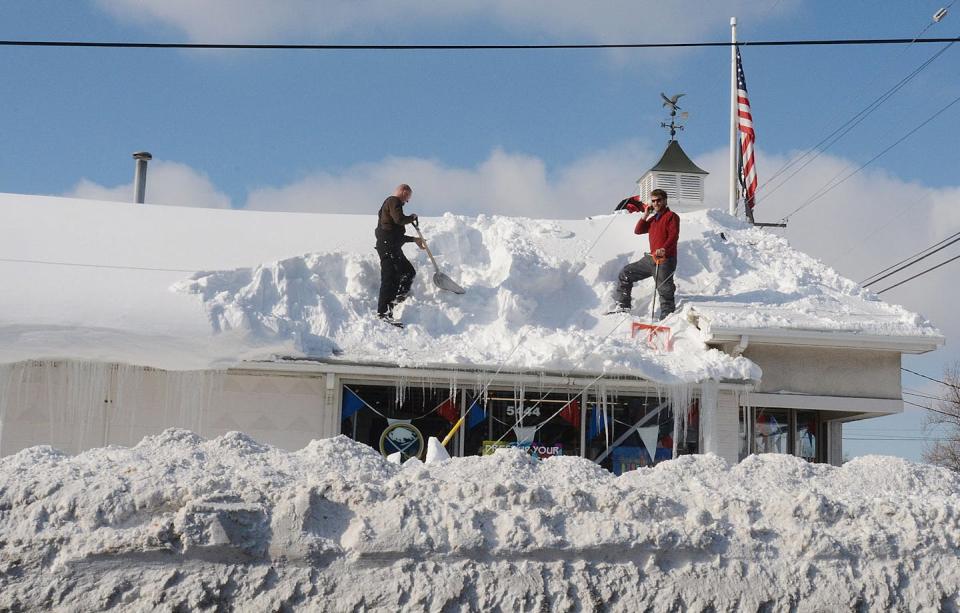 Two people remove knee-deep snow from a roof.