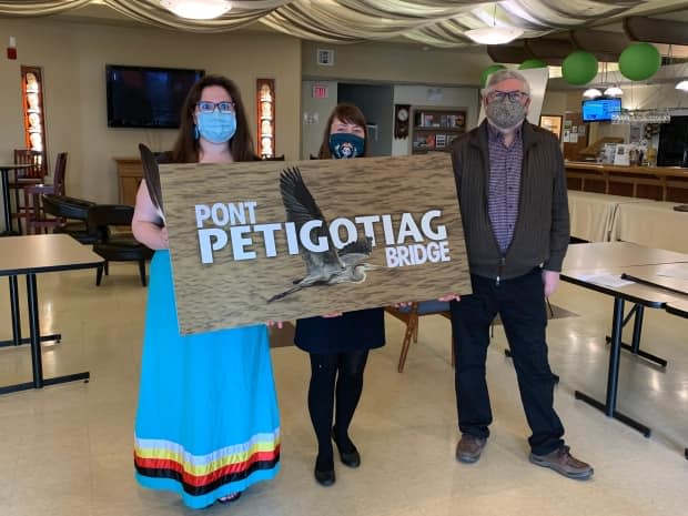 Michelle Knockwood, an Indigenous land conservation officer for the Fort Folly First Nation, Krysta Cowling, executive director of Petitcodiac Riverkeeper and Riverkeeper president Ronald Babin hold a sign with a proposed name for the new bridge between Moncton and Riverview. (Kate Letterick/CBC News - image credit)