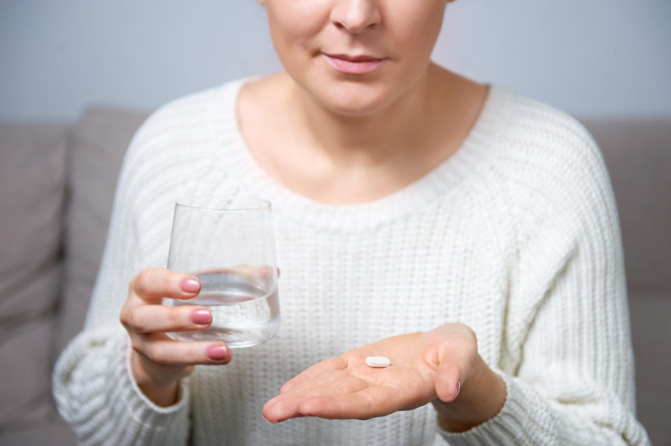 A woman holds a white pill in the palm of one hand and a glass of water in another