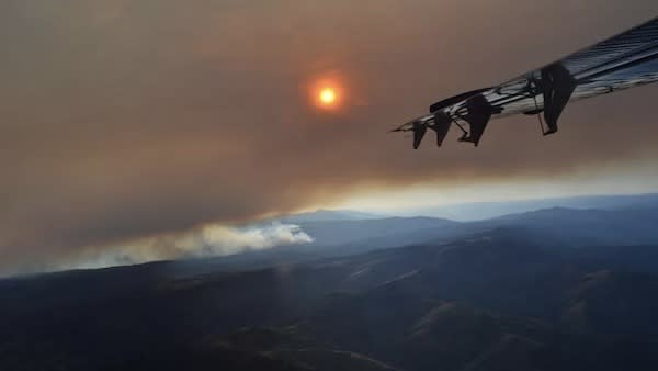 The wing of an airplane hangs over the right side of the image, a dimmed sun shines behind brown smoke above a dark, mountainous landscape. 