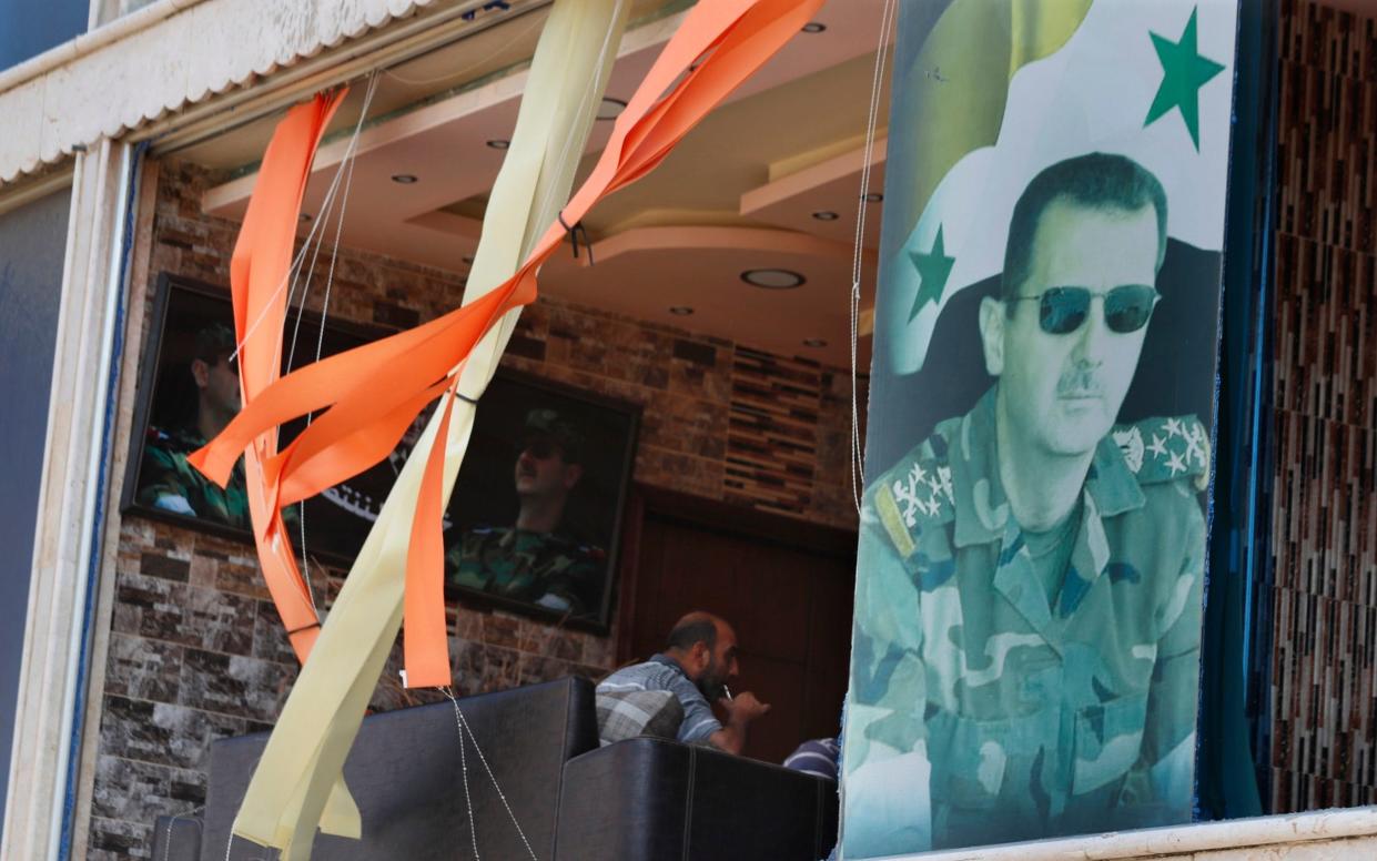 A man sits on a sofa in front of a blown-out window, next to the large portrait of Assad in military uniform and sunglasses