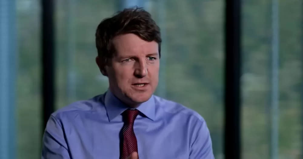 The image is a screen shot from an RTÉ Prime Time Documentary. It shows a headshot of a man wearing a blue shirt and wine coloured tie. He is sitting in front of a window with blurred out trees behind.