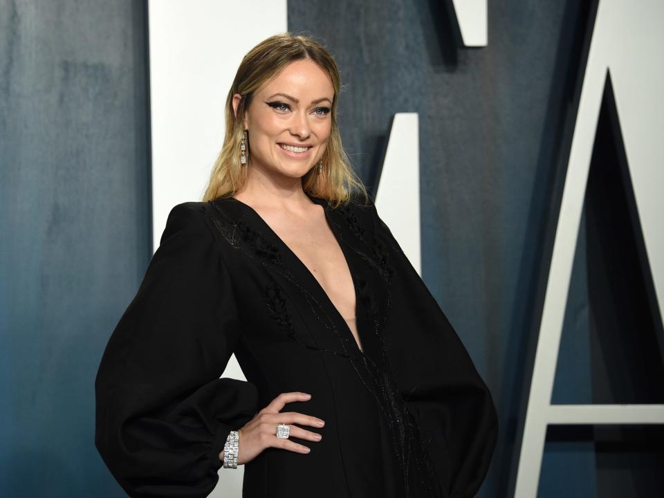 Wilde smiling in a wide-arm black gown in front of a Vanity Fair background.