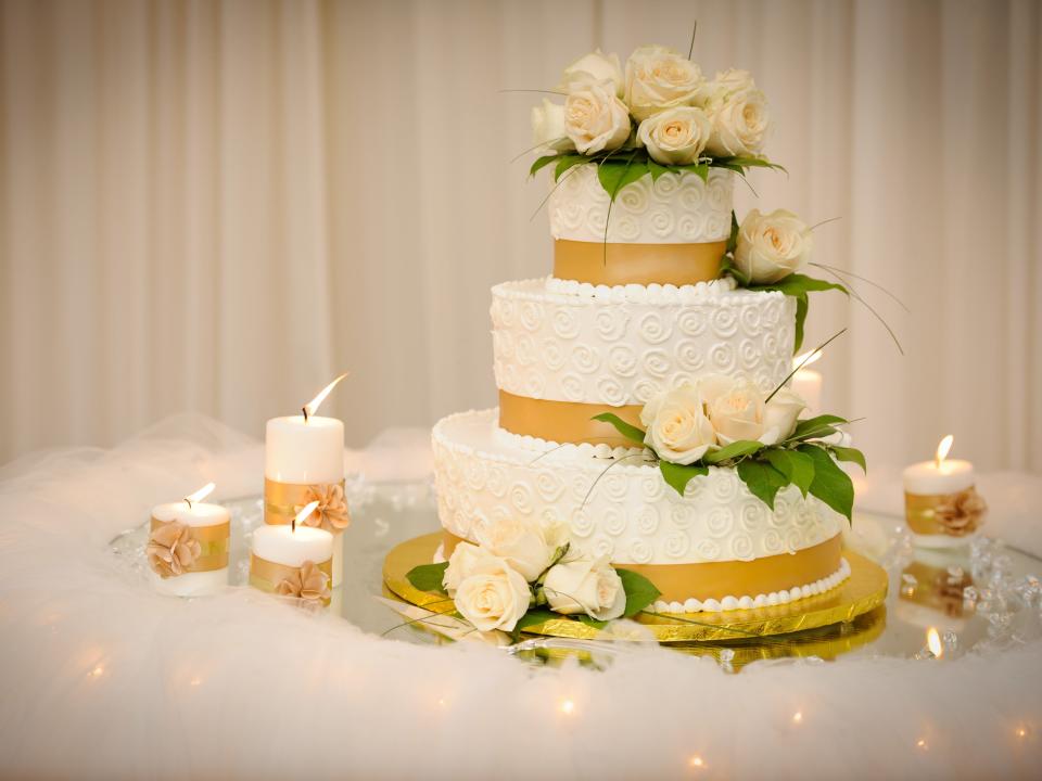 fancy tiered wedding cake with roses on it surrounded by candles on a white table