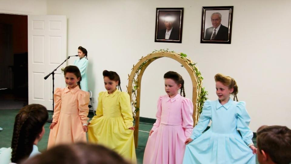 A group of very young girls dance in colorful, conservative dresses