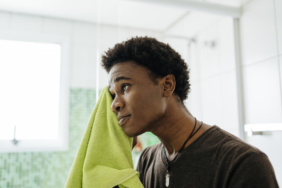 A person with short hair wipes their face with a towel in a bathroom. The background shows a window and mosaic tiles