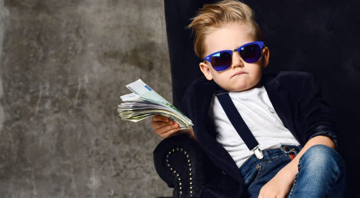 Una foto di un giovane ragazzo che indossa occhiali da sole, jeans, un blazer, una camicia bianca e reggicalze con in mano denaro di varie denominazioni e seduto su una sedia lussuosa.