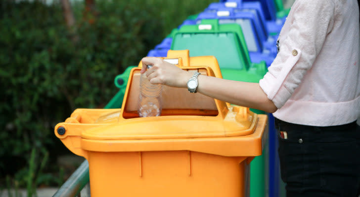 person depositing a plastic water bottle in a yellow plastic recycling bin. The bin is in a line-up of several other blue and green bins. dividend stocks for passive income