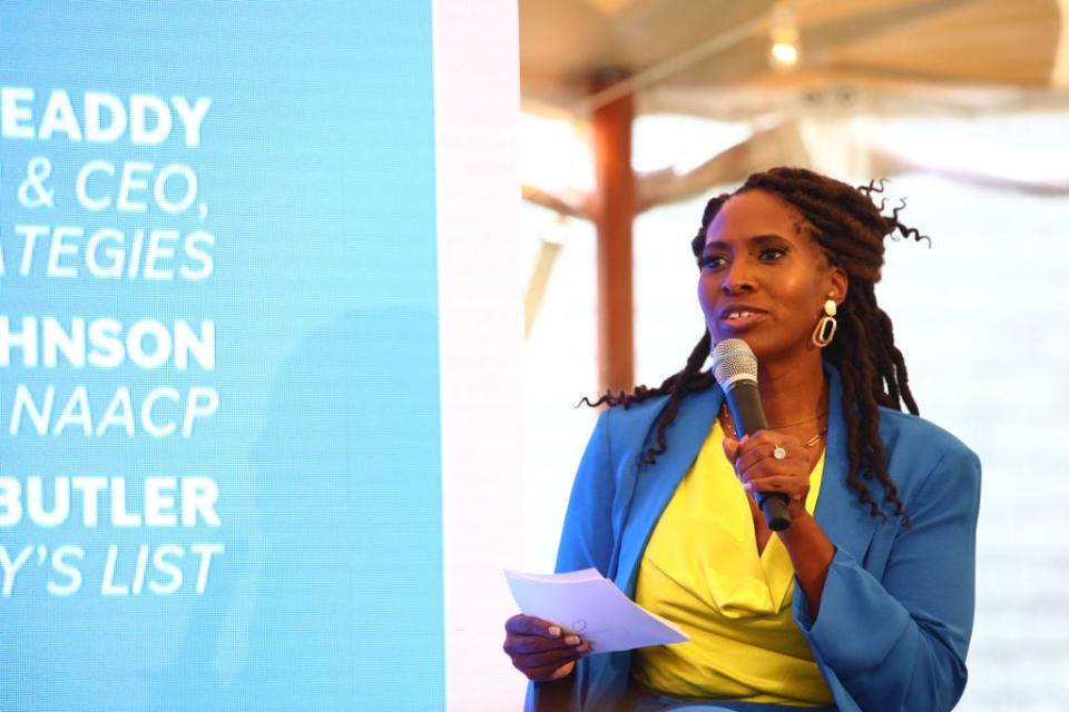 Jotaka Eaddy, wearing a yellow blouse and blue blazer, speaks into a microphone.