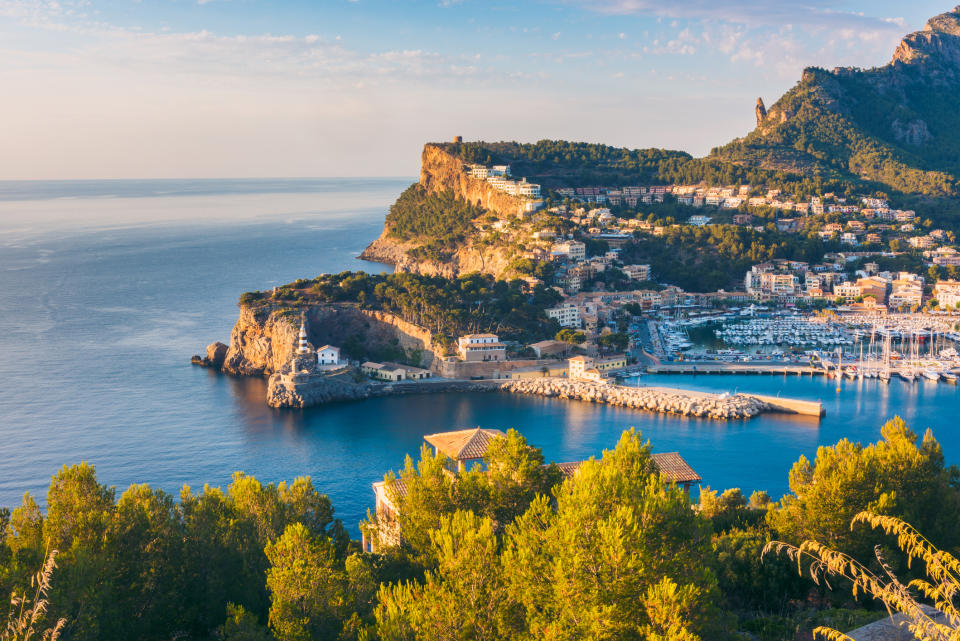 Scenic view of a coastal town with a marina, surrounded by lush green hills and cliffs, overlooking a calm blue sea