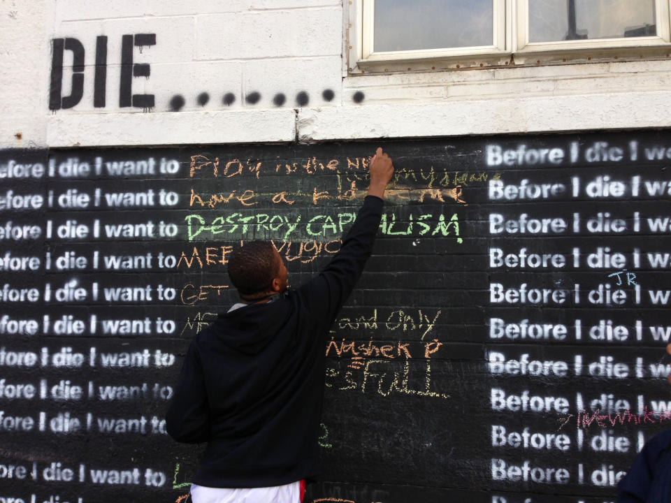 In this Nov. 7, 2013 photo, Nyquis Turner, 16, writes “Play in the NFL” on a wall in Syracuse, N.Y., that invites passers-by to complete the sentence: “Before I die, I want to...” The global phenomenon started in 2011, when artist Candy Chang created the first wall on an abandoned house in her New Orleans neighborhood. More than 400 walls have gone up in the United States and more than 60 other countries. (AP Photo/Carolyn Thompson)