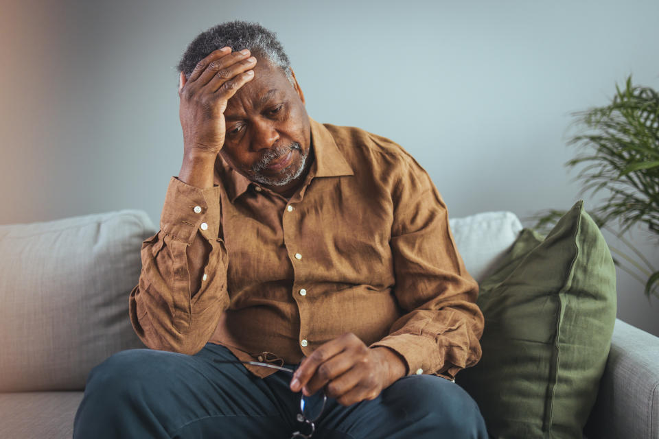 An older Black man sits on his sofa with a concerned expression on his face, as he leans his head into one hand