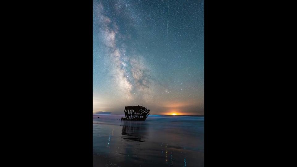 Bioluminescent tides glow beneath the Milky Way and a falling meteor from the Perseid shower near a shipwreck at Fort Stevens State Park in Oregon.