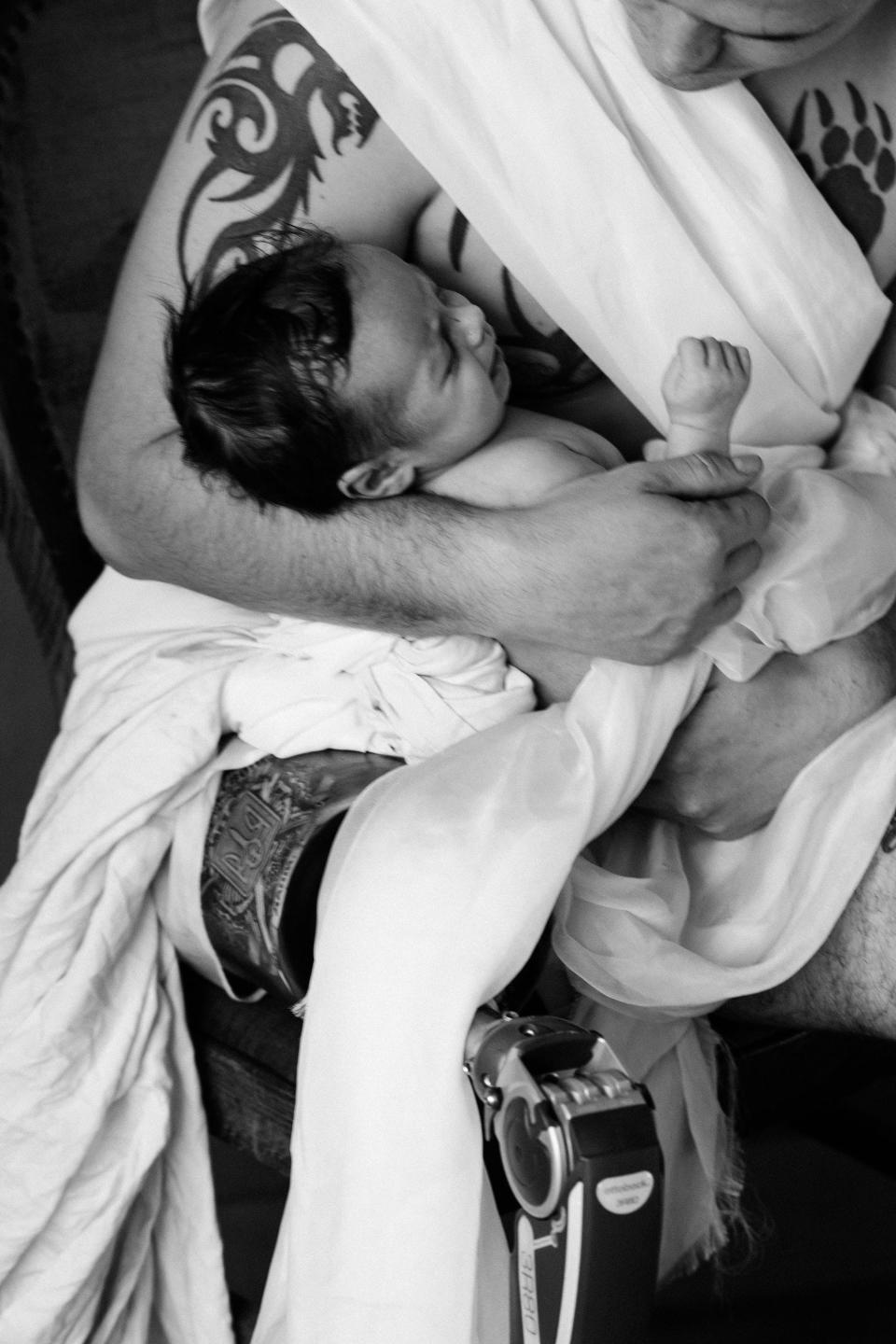 A closely-cropped black-and-white-photograph by Ukrainian photographer Marta Syrko, showing soldier Serhii and his son. Serhii is seated and unclothed except for long bands of white cloth, under which his tattoos can be seen. He looks down at his baby son in his arms. In the lower half of the picture, his prosthetic lower leg is visible.