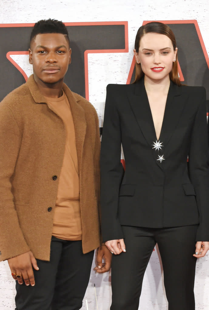 John Boyega and Daisy Ridley pose together on a red carpet. John wears a brown coat over a tan shirt, and Daisy wears a black blazer with starburst brooches