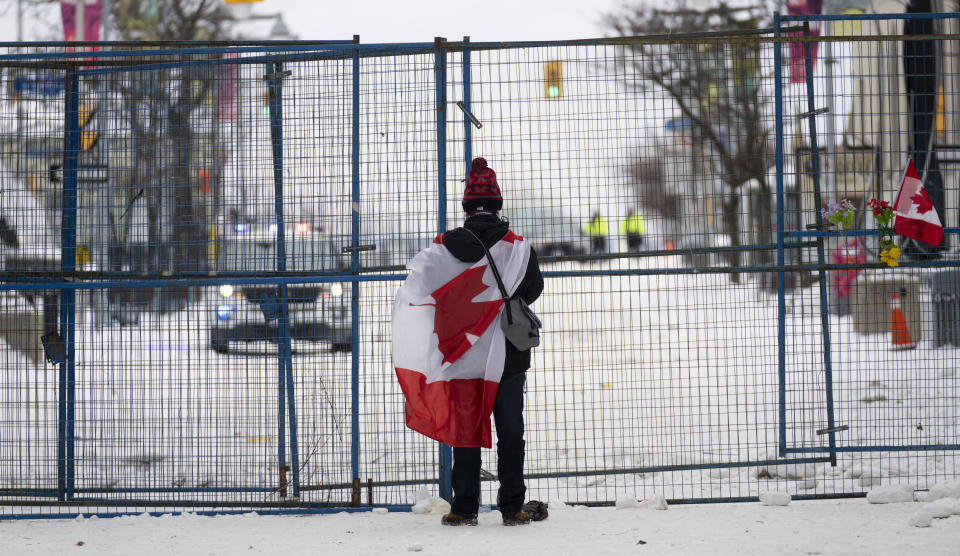 A lone protester stands draped in the Canadian flag at a temporary fence controlling access to streets near Parliament, in Ottawa, Sunday, Feb. 20, 2022. (Adrian Wyld/The Canadian Press via AP)