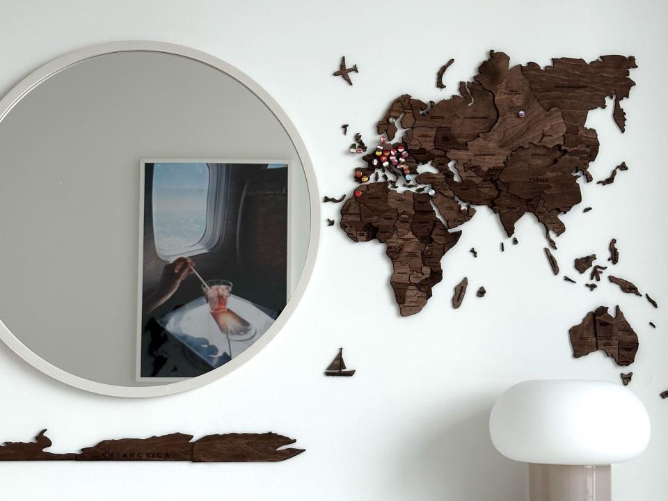 On the left is a mirror with a photo taped to it of a hand stirring a drink at a window seat on a plane. To the right is a wooden map with pins in it.