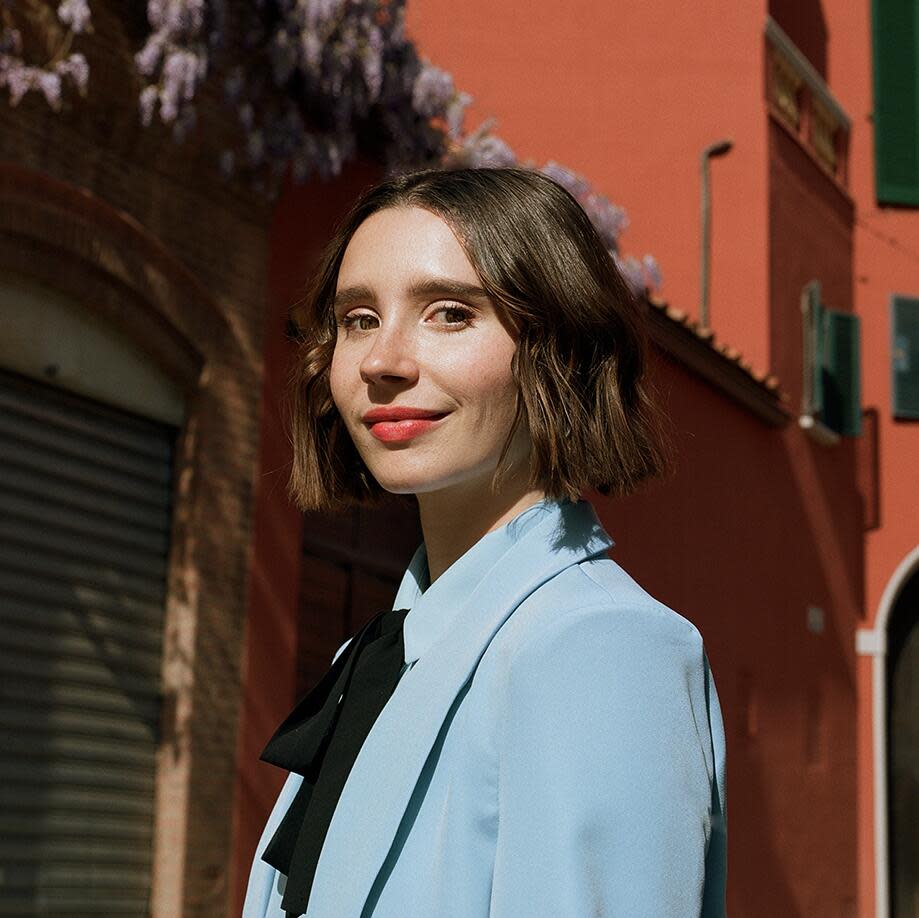 A smiling woman with long brown hair, wearing a light blue blazer, stands outside a building