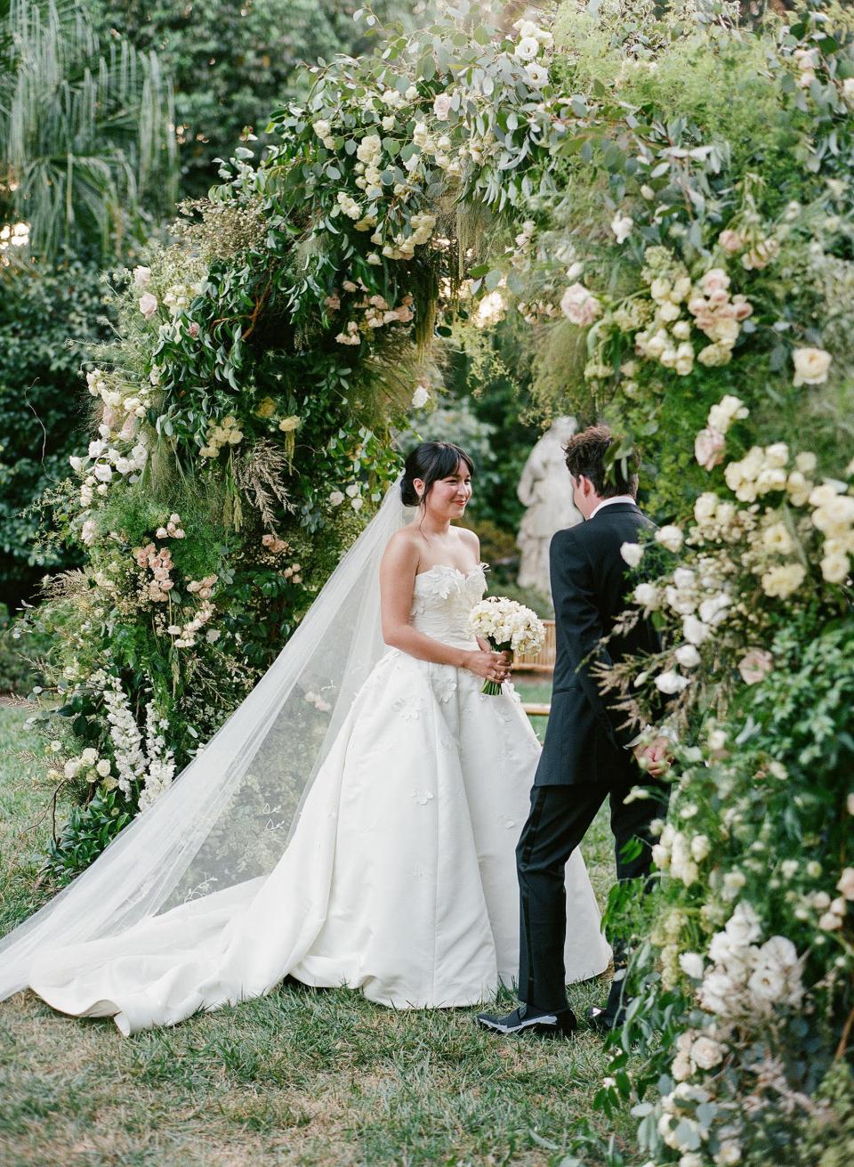 A bride looks at her groom during their wedding ceremony under a floral archway.