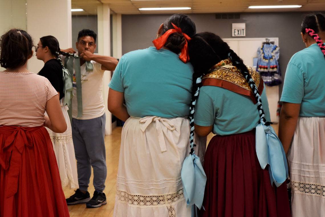 Maritza Estrada (16, with a red bow) and Leylie Garduno (17, with blue ribbon braids) embrace while their dance director, Hugo Martínez, demonstrates how tight braid bows should be with Andrea Felix-Gastelum (17, with green ribbon braids) during their Grupo Folklórico Tangu Yuu rehearsal on Sept. 20, 2022 in Fresno.