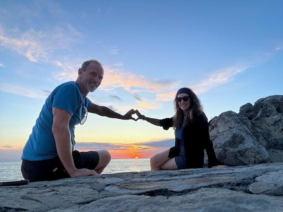 Dorine Olive and her partner, Chuck, making a heart sign in front of a sunset by a beach.