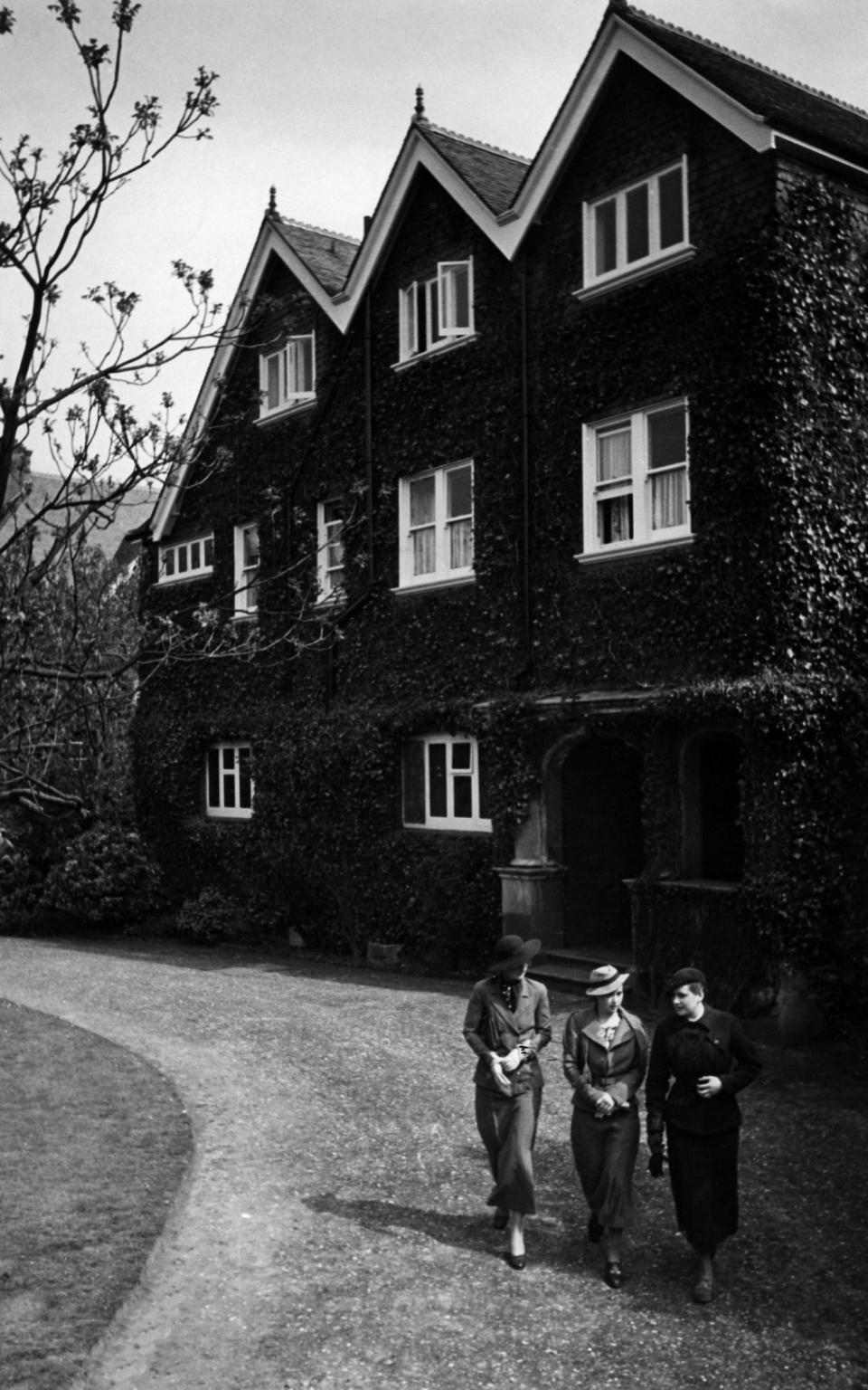 Black and white photo of a dark brick building with three young women walking down its driveway - ullstein bild