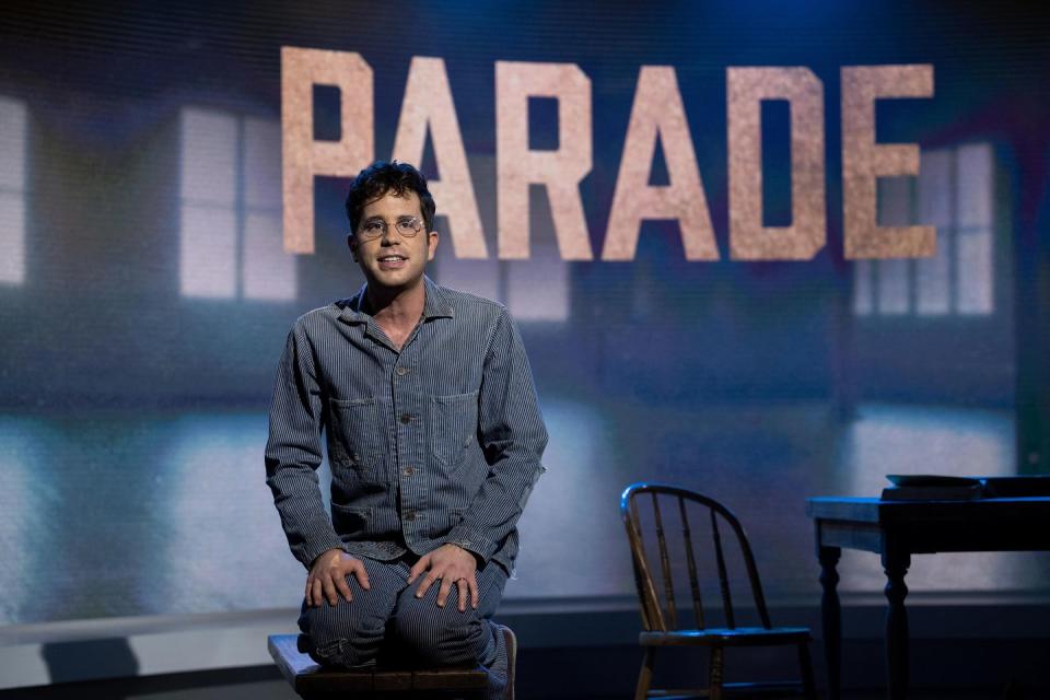 ben platt wearing a blue denim shirt and jeans and glasses, kneeling in front of a darkened wall with the words parade on it