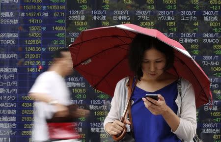 A woman holding an umbrella looks at her cellphone in front of an electronic board showing the various stock prices outside a brokerage in Tokyo August 8, 2014. REUTERS/Yuya Shino