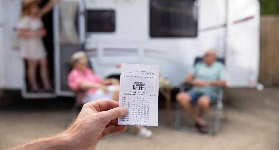 Lotto ticket being held in front of caravan with two people in chairs. 