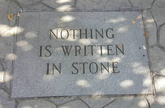 The image shows a stone plaque with the phrase "NOTHING IS WRITTEN IN STONE" engraved on it, surrounded by leaves and sunlight