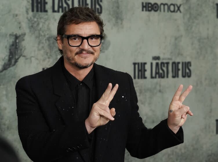 Pedro Pascal in black glasses and a black suit holding up peace signs with both his hands