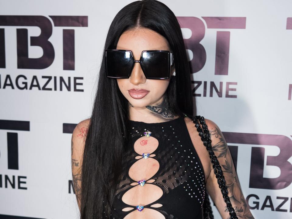bhad bhabie on a red carpet. she's wearing black fishnet tights and a black unitard, that has openings running from her neck to her midriff. her hair is worn long, and she's wearing large, blocky sunglasses