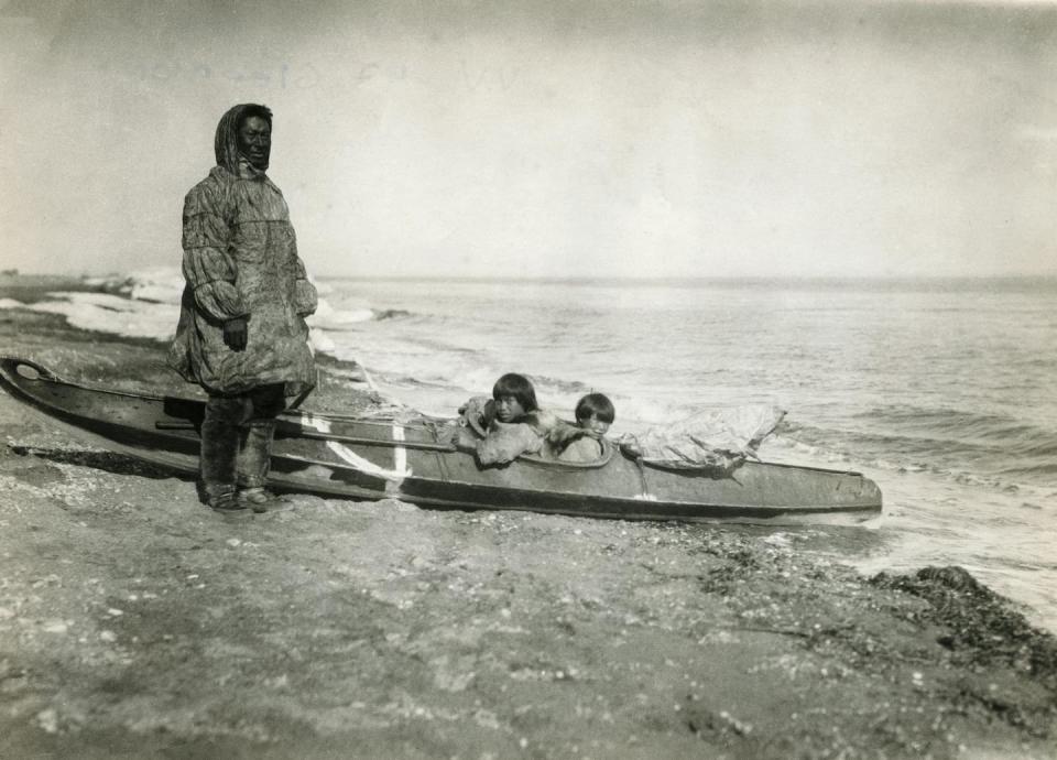 man in traditional clothes stands next to canoe with two children in it on the shoreline