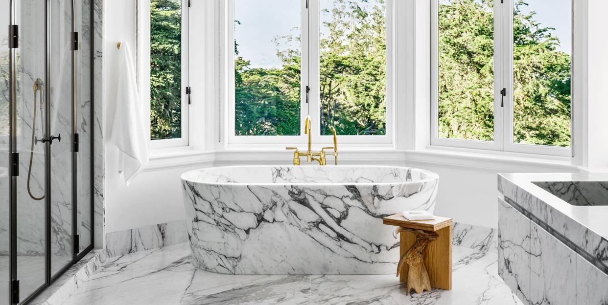 rounded bathroom with double windows in an arc at the back with a standalone marble tub at center and a floating marble vanity and a large bat like light fixture at center and a glass shower at left