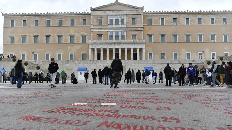 La gente pasa junto a los nombres de las víctimas de la colisión de trenes del año pasado pintados en una acera frente al parlamento, en Atenas, Grecia, el 28 de feb