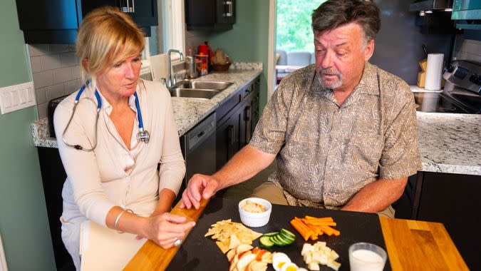 A senior man talking with a visiting nutritionist at home, A selection of healthy food choices are on display.