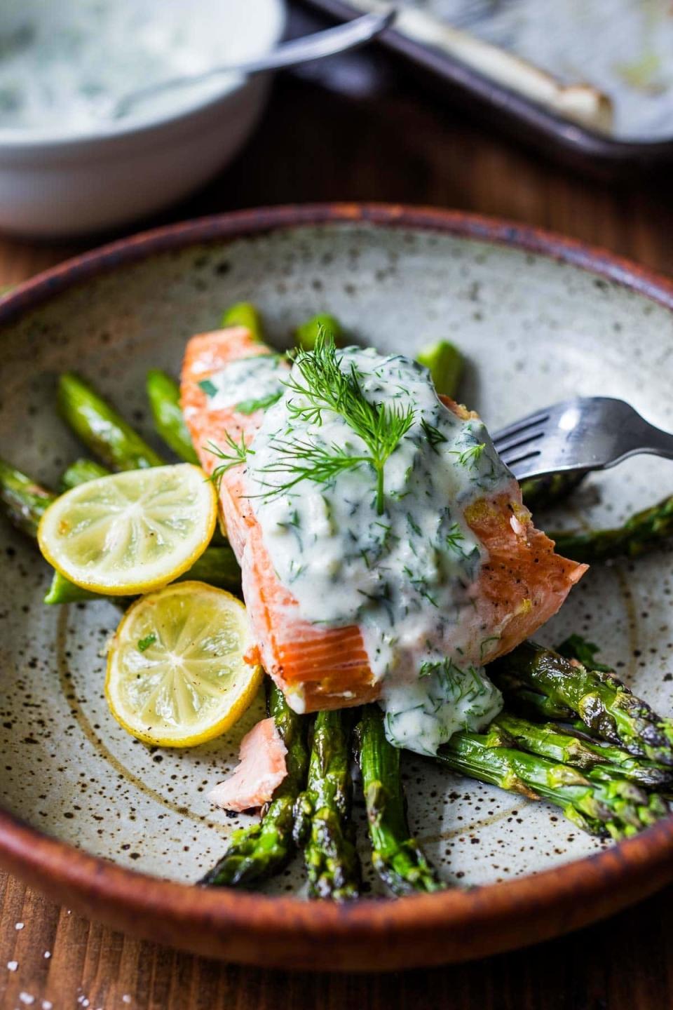 Grilled salmon with asparagus topped with dill sauce and lemon slices on a plate with a fork