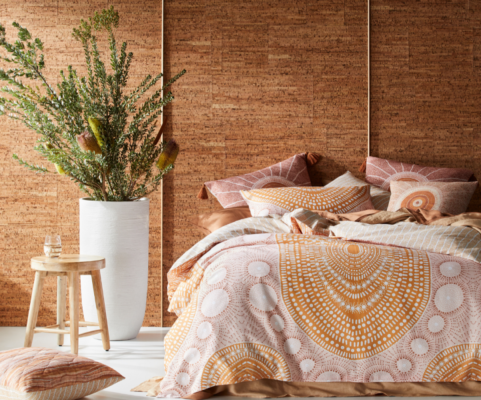 Image of a bed dressed with an indigenous orange and pink pattern quilt cover in front of a brown wall with a native plant to the right.