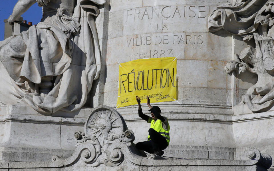 A protestor in a yellow vest sits under a banner which reads 'revolution' at the Place de Republique, during a yellow vest demonstration in Paris, Saturday, April 20, 2019. French yellow vest protesters are marching anew to remind the government that rebuilding the fire-ravaged Notre Dame Cathedral isn't the only problem the nation needs to solve. (AP Photo/Michel Euler)