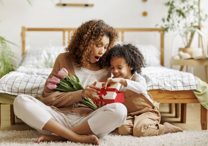 Cheerful african american family mom opens a gift box with her son and holding bouquet  of flowers while resting on floor by bed during holiday celebration mothers day at home